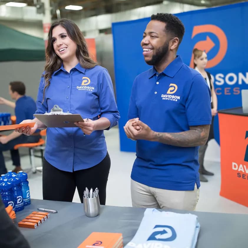 Two coworkers wearing coordinating branded uniforms, talk to a prospective client at their booth featuring promotional giveaways and custom signage.