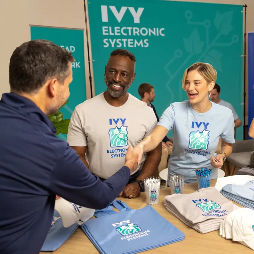 Coworkers wearing coordinating branded t-shirts greet a prospective client at their tradeshow booth, featuring custom signage and promotional giveaways.