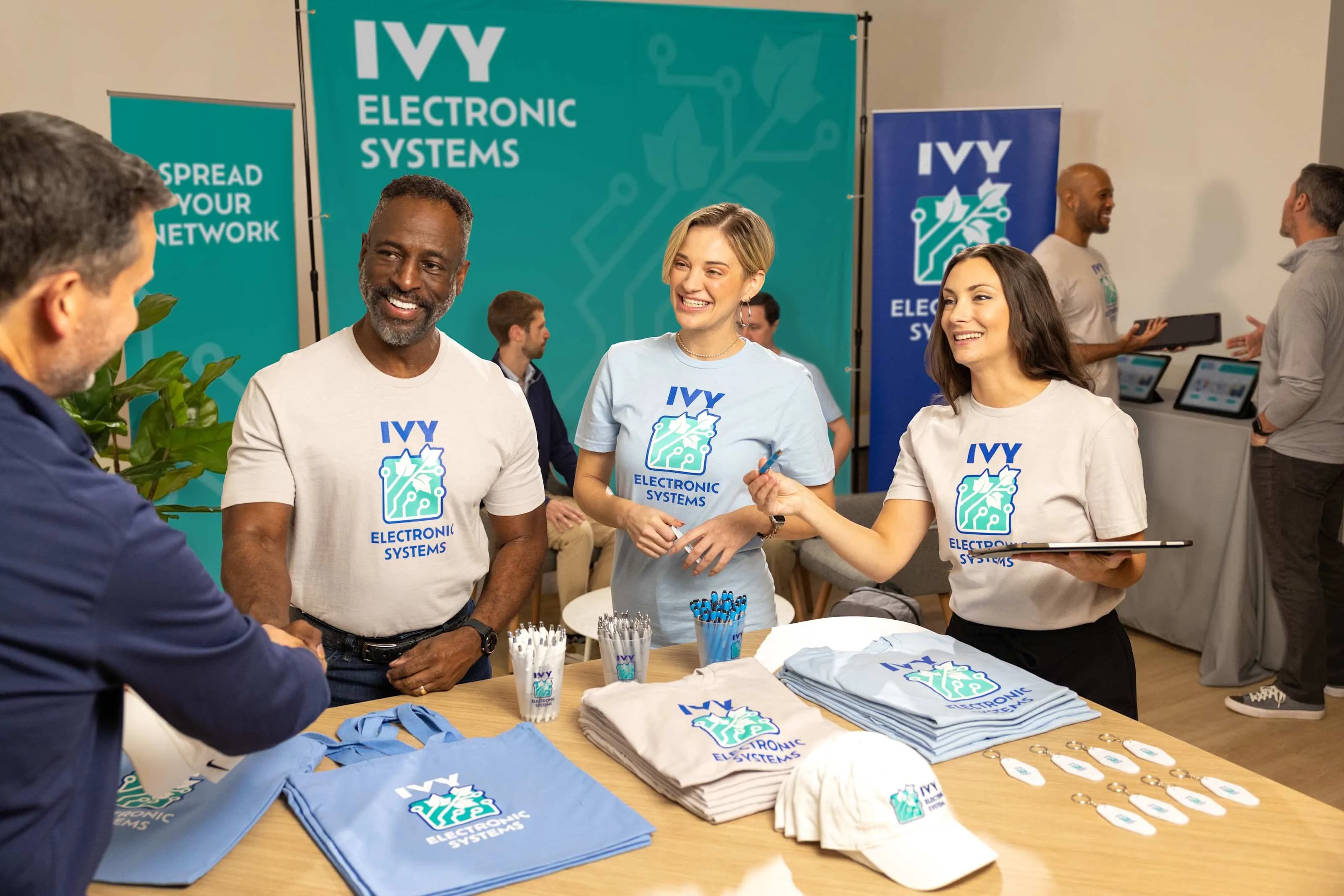 Three coworkers at a tradeshow, wear matching logo t-shirts behind a table display of branded promotional giveaways.