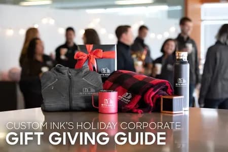 Branded corporate gifts including custom logo apparel, mugs, and blankets displayed on a table with coworkers in the background.